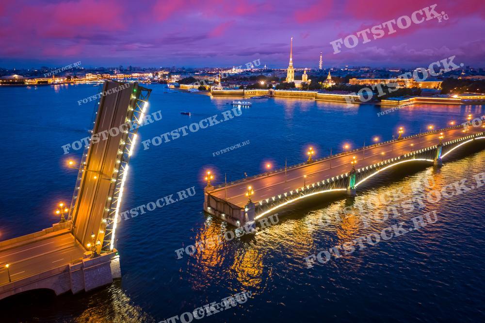 На фотографии изображен разведенный Троицкий мост в Санкт-Петербурге ночью. Мост ярко освещен, создавая контраст с темным небом и отражаясь в воде Невы. На заднем плане видна Петропавловская крепость с золотым шпилем Петропавловского собора. Красочное небо добавляет атмосферу волшебства ночному Петербургу.